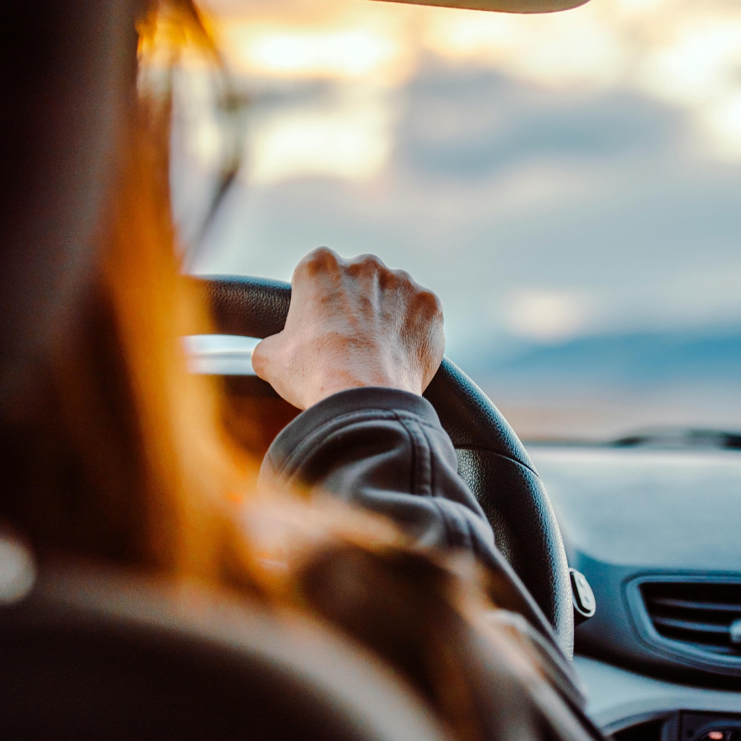 A person's hands gripping a steering wheel from the driver's perspective, looking out at a road with a cloudy sky in the background, conveying the act of driving.