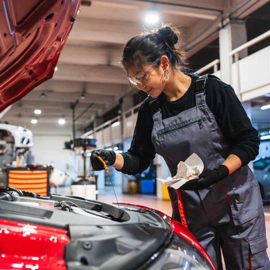 A smiling mechanic in grey overalls and safety glasses checking the oil of a red car in a garage, holding a dipstick and a white rag.