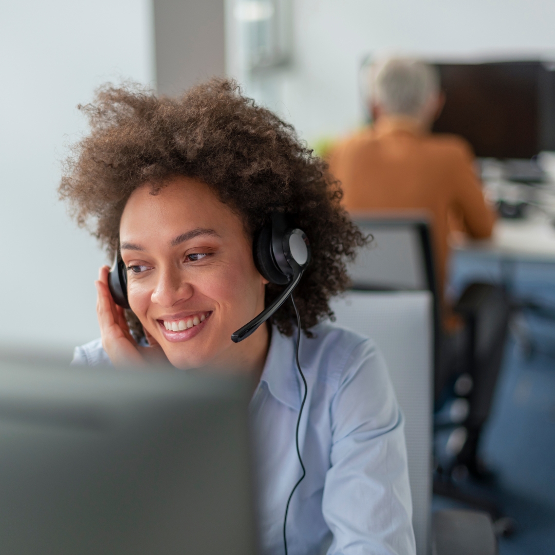 A smiling woman with curly hair wearing a headset and looking at a computer screen, likely working in a customer service or call center environment.