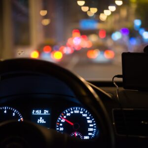 A view from the driver's seat at night, showing the illuminated dashboard speedometer and the blurry bokeh lights of traffic outside the windshield.