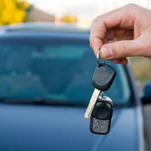 A close-up of a person's hand holding a car key and key fob against a blurred background of a dark blue car hood.